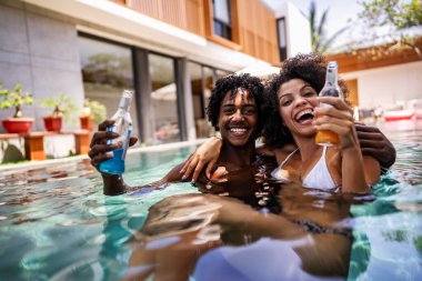 Genç bir çift Afrikalı lüks bir havuz başında güneşli bir günün tadını çıkarıyor, renkli içkilerle gülüyor ve kadeh kaldırıyor. Üzerlerinde mayo var, etrafı tropikal ve rahatlatıcı bir atmosferle çevrili.