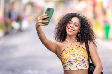 Genç ve güzel Afrikalı Amerikalı kadın mutlu bir gülümsemeyle gülümsüyor ve akıllı telefonuyla selfie çekiyor.  