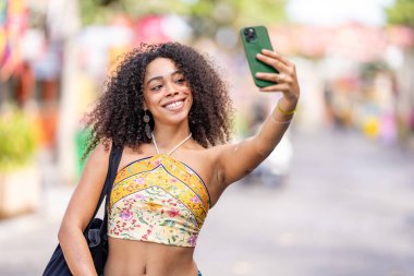 Genç ve güzel Afrikalı Amerikalı kadın mutlu bir gülümsemeyle gülümsüyor ve akıllı telefonuyla selfie çekiyor.  