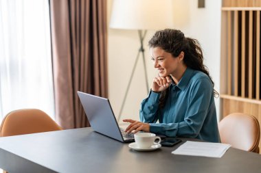Genç bir kadın kahve içerken dizüstü bilgisayarda çalışıyor.