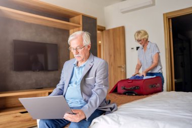 Karısı bavulları açarken yaşlı işadamı otel odasında dizüstü bilgisayar kullanıyor.