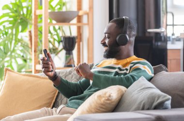 Kulaklıklı mutlu Afro-Amerikalı adam cep telefonuyla müzik dinliyor, kanepede dinleniyor. 