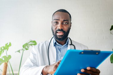 Beyaz önlüklü genç Afrikalı doktor elinde haplar ve kontrol listesi tutuyor. 