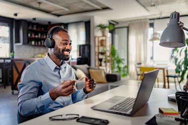 Kulaklıklı genç erkek girişimci ev ofisinde otururken dizüstü bilgisayar kullanıyor.
