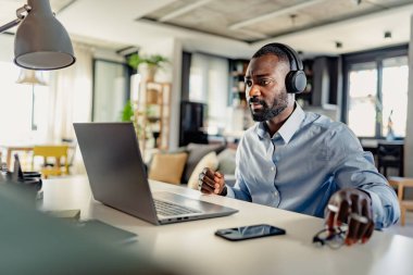 Kulaklıklı genç erkek girişimci ev ofisinde otururken dizüstü bilgisayar kullanıyor.