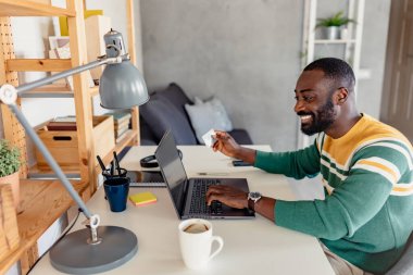 Genç Afrikalı gülümseyen ve elinde kredi kartı tutan bir adam online alışveriş yapıyor. 