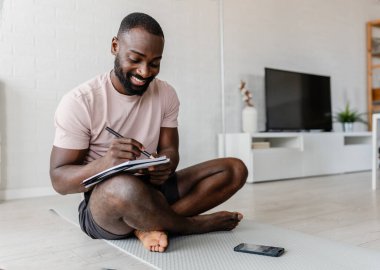 Gülümseyen adam yere oturmuş deftere yazıyor. 