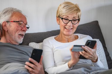 Olgun bir çift yatakta yatarken online alışveriş yapıyor. 