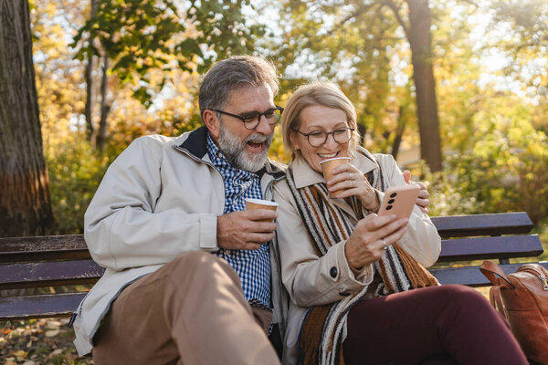 mature couple with smartphone sitting on the bench in park 