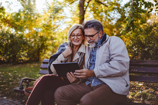 senior couple with tablet sitting on bench in park 