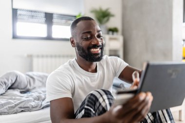 Genç siyah adam tablet kullanırken gülümsüyor ve online alışveriş yapıyor. 