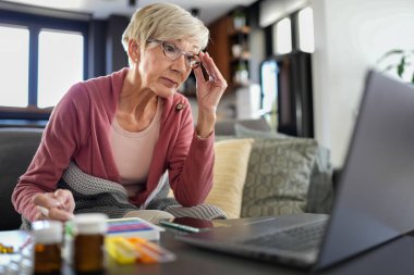 Masada dizüstü bilgisayar kullanarak oturan gözlüklü yaşlı kadın.