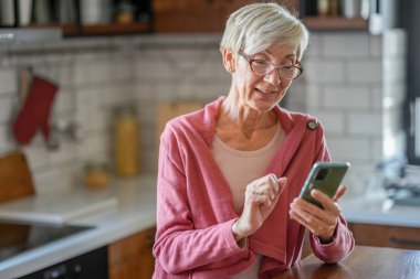 Gülümseyen yaşlı kadın mutfak tezgahında akıllı telefon kullanıyor.