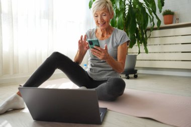 Evde dizüstü bilgisayar ve akıllı telefon kullanarak oturan orta yaşlı bir kadın. 