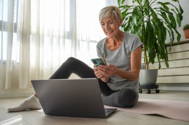 Evde dizüstü bilgisayar ve akıllı telefon kullanarak oturan orta yaşlı bir kadın. 