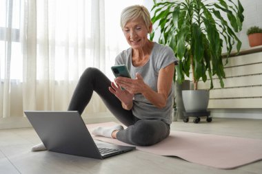 Evde dizüstü bilgisayar ve akıllı telefon kullanarak oturan orta yaşlı bir kadın. 
