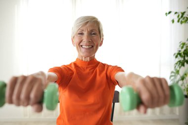 Gülümseyen olgun kadın evde egzersiz yaparken dambılları kaldırıyor.