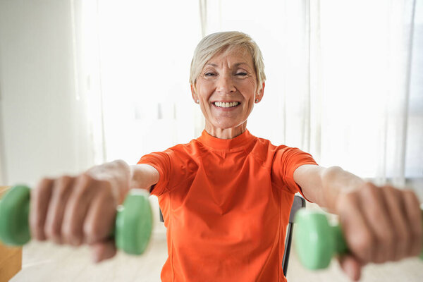 Улыбающаяся взрослая женщина поднимает гантели, упражняясь дома