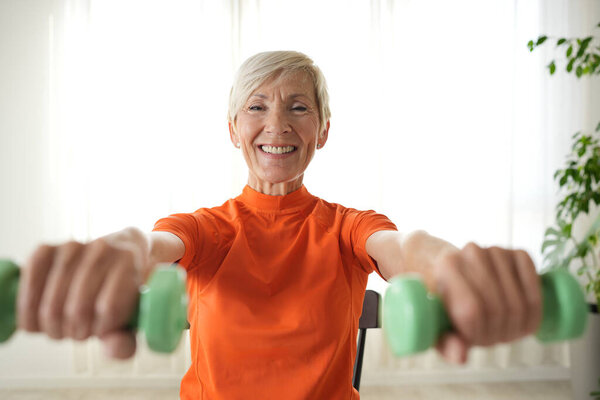 Улыбающаяся взрослая женщина поднимает гантели, упражняясь дома