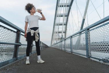Genç kadın modern köprüde dururken su içiyor.