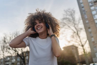 Gülümseyen genç bir kadın gün batımında açık havada müzik dinliyor.