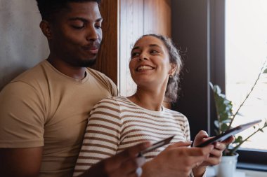 Pencerenin yanında akıllı telefonlar kullanan gülümseyen çift.