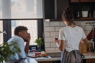 Şarap kadehi tutan kadın mutfakta bir adamla konuşuyor.