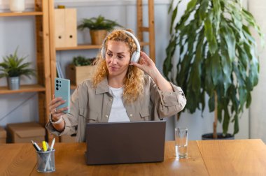 Kulaklık takan genç bir kadın akıllı telefon kullanarak masada oturuyor.