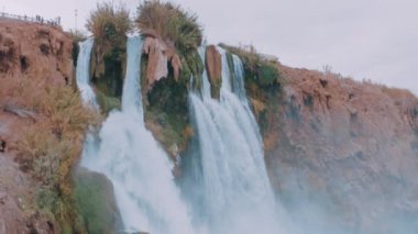 Güzel fırtınalı şelale. Kayanın üzerinde yoğun bitki örtüsü var. Mavi göl ve kayadaki otel. İHA video 4K görüntüsü. Yüksek kalite 4k görüntü