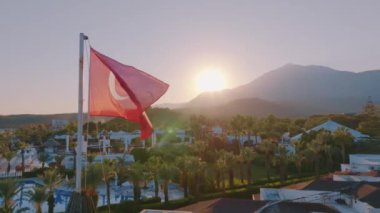 Çatıda Türk bayrağı dalgalanıyor. Mavi yüzme havuzu, arka planda palmiye ağaçları ve dağlar. Gün batımı. Yüksek kalite 4k görüntü