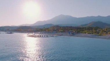 Gün batımında deniz manzarası. İskele denize açılır. Kumsal, şemsiyeler, dinlenme yerleri. Dinlenmek için bir yer. Arka planda dağlar. Hava aracı video görüntüleri. Yüksek kalite 4k görüntü