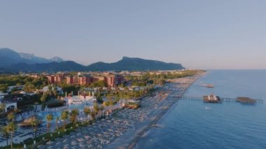 Kıyı şeridi üzerinde uçuş. Plaj, oteller, palmiye ağaçları, şezlonglar, iplik şemsiyeleri. Tepeler ve dağlar arka planda. Yüksek kalite 4k görüntü