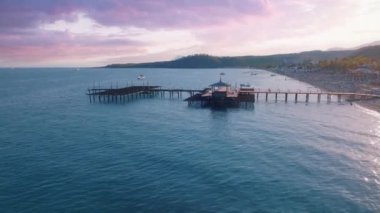 Gün batımında deniz manzarası. İskele denize açılır. Kumsal, şemsiyeler, dinlenme yerleri. Dinlenmek için bir yer. Arka planda dağlar. Hava aracı video görüntüleri. Yüksek kalite 4k görüntü