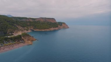 Denizde büyük bir kayalık adanın üzerinden uçmak. Kumsalda güneşli şezlonglar ve güneşlikler. Tekne kalkıyor. Hava aracı video görüntüleri. Yüksek kalite 4k görüntü