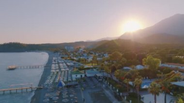 Hava aracı video görüntüleri. Gün batımında sahil şeridine uçun. Kumsal, denizde iskele, oteller ve palmiye ağaçları. Tepeler ve dağlar arka planda. Yüksek kalite 4k görüntü