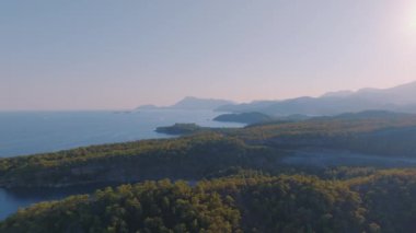 Hava aracı video görüntüleri. Gün batımında deniz manzarası. Kıyıda mavi deniz ve yeşil ağaçlar. Arka planda dağlar. Yüksek kalite 4k görüntü