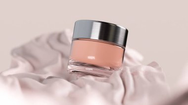 Gümüş kapaklı bir kozmetik krem kavanozunun 3 boyutlu görüntüsü zarif tasarımını sergileyen pembe kumaşın üzerine güzelce yerleştirilir.