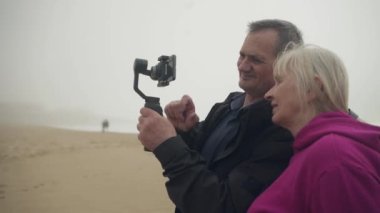 Çekici son sınıf beyaz beyaz çift cep telefonundan görüntüler izliyor ve sahilde gülüyorlar. Adam elinde bir telefon zili tutuyor. Yüksek kalite 4K görüntü