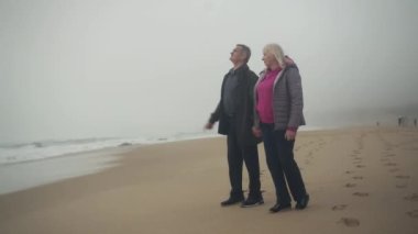 Arka planda fırtınalı okyanusla sahilde el ele tutuşarak yürüyen son sınıf beyaz bir çift. Yağmurluk giyip neşeyle sohbet ediyorlar. Geniş açı. Yüksek kalite 4K görüntü