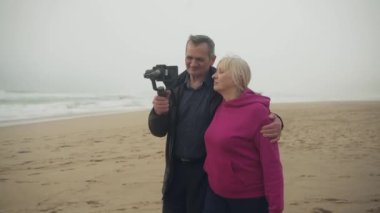 Çekici, neşeli, beyaz, son sınıf öğrencisi çift, fırtınalı bir havada plajda telefon sinyali kullanarak sosyal medya için bir video çekiyor. Orta boy. Yüksek kalite 4K görüntü