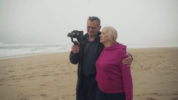 魅力的な陽気な白人白人のシニアカップルは 電話ジンバルを使用して荒天時にビーチでソーシャルメディアのビデオを撮影します 中発だ 高品質4K映像 — ストック動画
