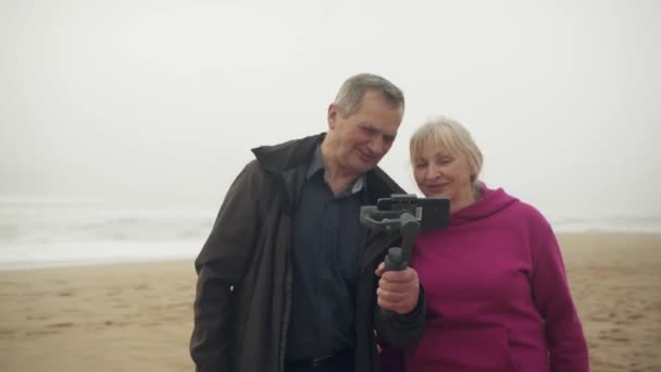 迷人的资深白人白人夫妇一边看手机上的镜头 一边在海滩上大笑 男人手里拿着话筒 高质量的4K镜头 — 图库视频影像