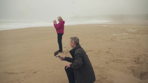 Starszy Biały Mężczyzna Filmujący Plaży Pomocą Gimbalu Kobieta Tle Medytuje — Wideo stockowe