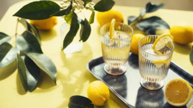 Su ve limonlu bardaklar. Güneşli bir günde limon ve çiçeklerle süslenmiş bir masada sağlıklı içecekleri tazelemek. Yüksek kalite 4K görüntü