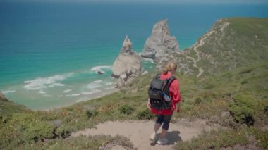 Beyaz kadın, kırmızı tişört ve siyah fotoğraf sırt çantası giymiş Atlantik kıyısı boyunca manzaralı uçurumlarda yürüyüş yapıyor. Portekiz 'in Cascais-Sintra Doğal Parkı' ndaki Ursa Sahili manzaralı. 