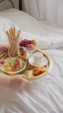 Yatağa beyaz çarşaf ile kahvaltı tepsisi koymak. Sağlıklı bir avokado, somon tostu, taze meyve ve bir fincan kahve içeren altın tepsi. Dikey video, 4K