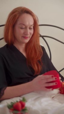 Rahat bir yatakta beyaz saçlı kızıl bir kadın kalp şeklinde bir hediye kutusunu açıyor. Kalp desenli pijama giyiyor. Dikey video