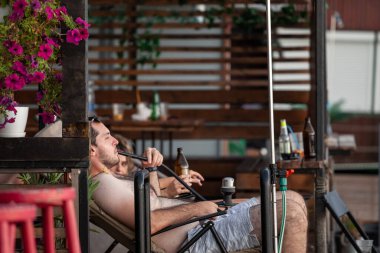 BELGRAD, SERBIA - 31 Temmuz 2021: Adam mayo üzerinde dinleniyor, nargile tütünü içiyor ve bira içiyor