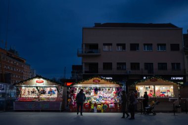 NOVI SAD, SERBIA - 4 ARALIK 2022: Novi Sad Noel pazarının butiklerinde seçici bulanıklık gece trg slobode meydanındaki el sanatları kulübelerinden ve tezgahlardan bir şeyler satın alan insanlar.