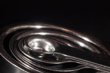 Stüdyodaki siyah bir arkaplanda izole edilmiş ölçüm bardakları gibi çeşitli miktarlarda, kaşık, çay kaşığı, çeşitli miktarlarda paslanmaz çelikten yapılmış makro bir karede seçici bulanıklık..
